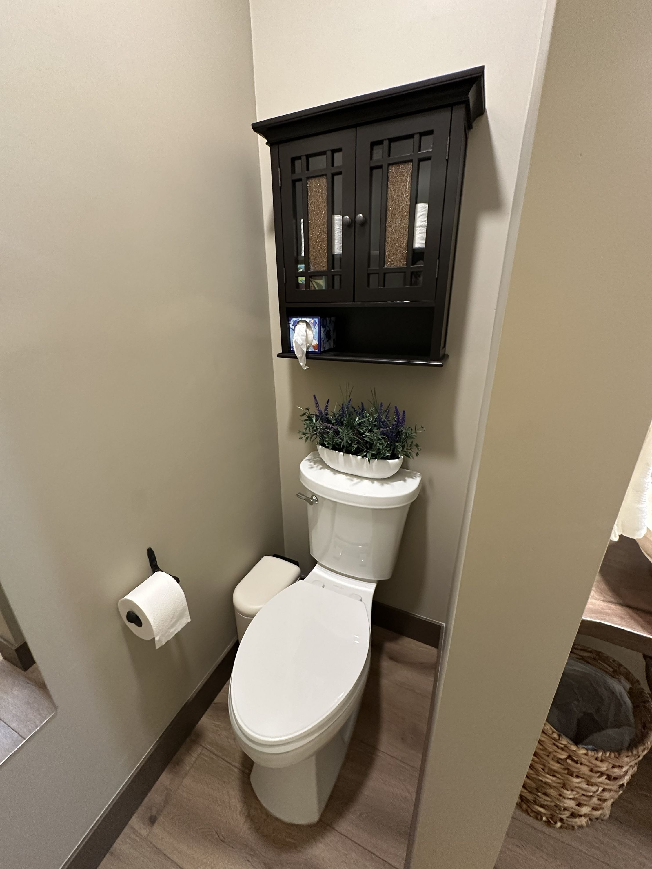 A bathroom with a toilet, a toilet paper holder, a shelf with a plant, and a basket.