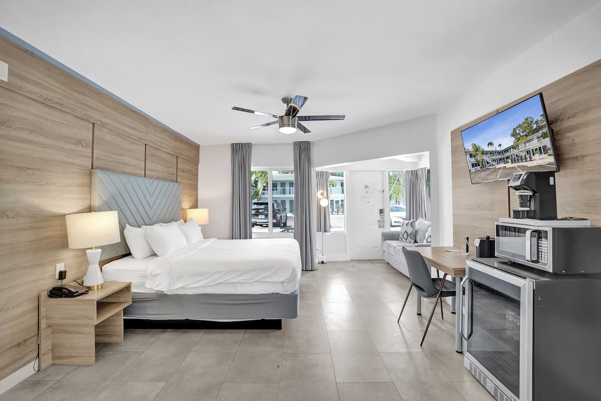 A modern hotel room with a bed, desk, TV, and kitchenette.