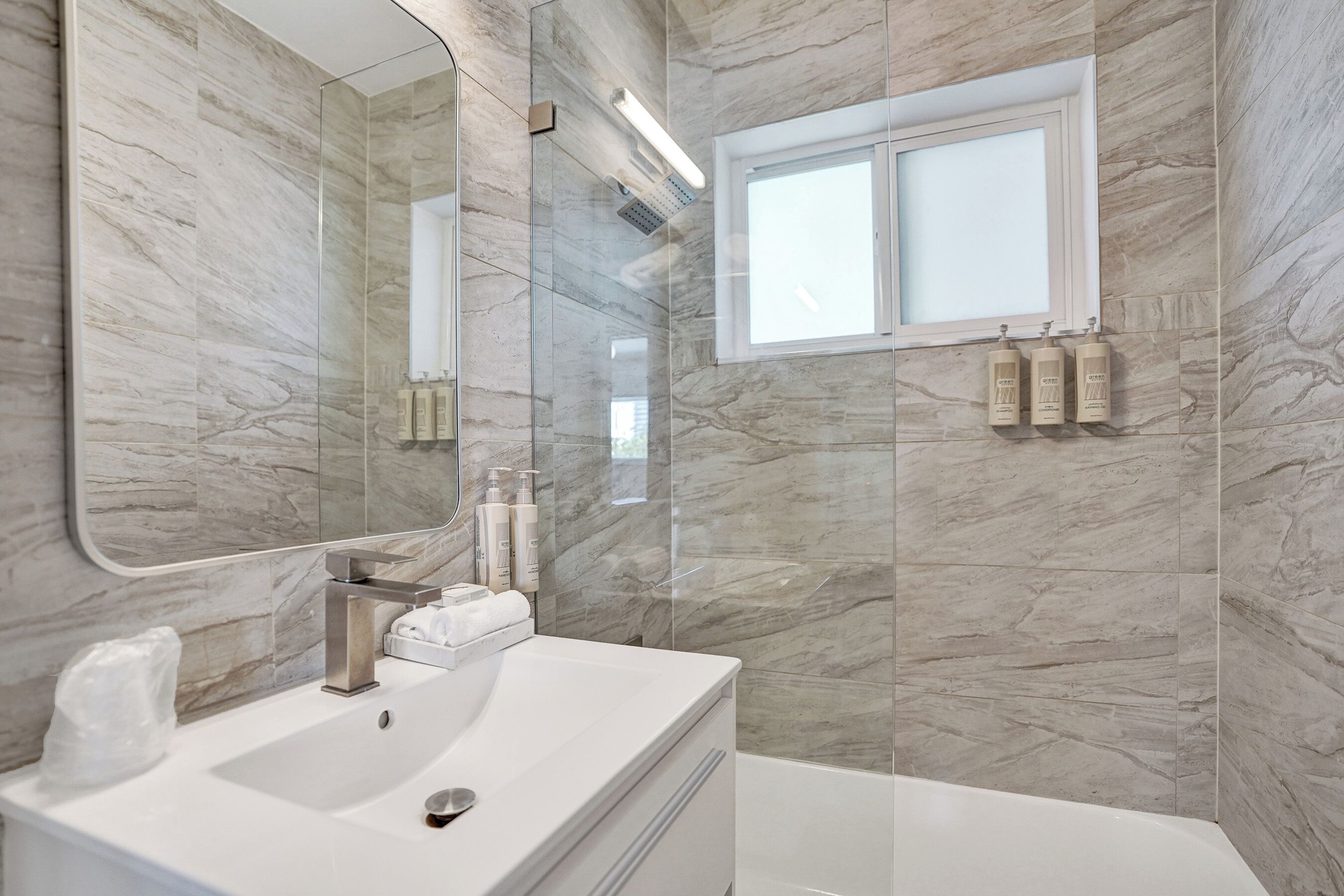 Un baño moderno con pared de mármol, un espejo grande, un lavamanos y una ventana.