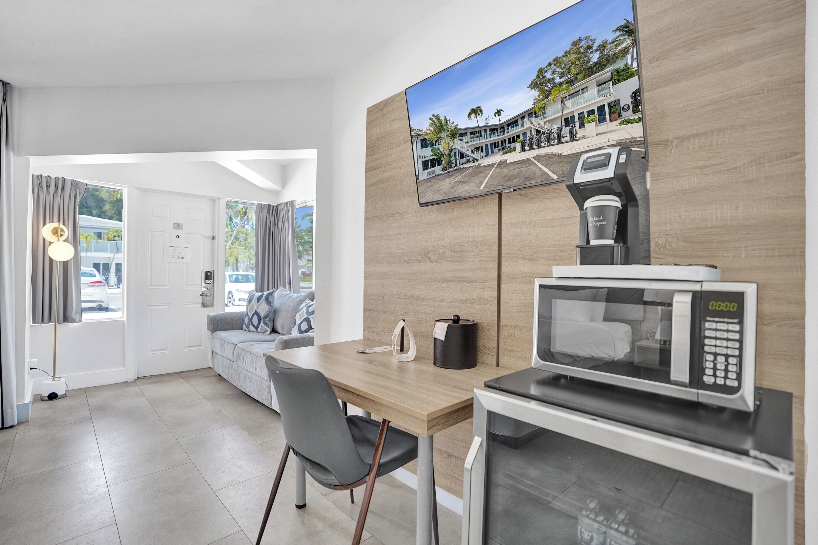 Habitación de hotel moderna con escritorio de madera, microondas, cafetera, televisor de pantalla plana y sofá con vistas al exterior.