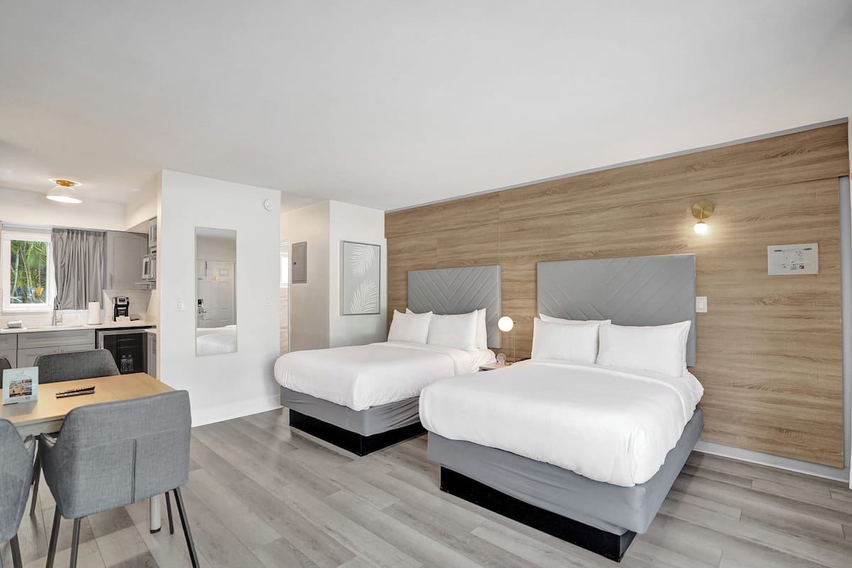 A modern hotel room with two beds, a dining area, and a kitchenette.