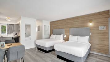 A modern hotel room with two beds, a dining area, and a kitchenette.