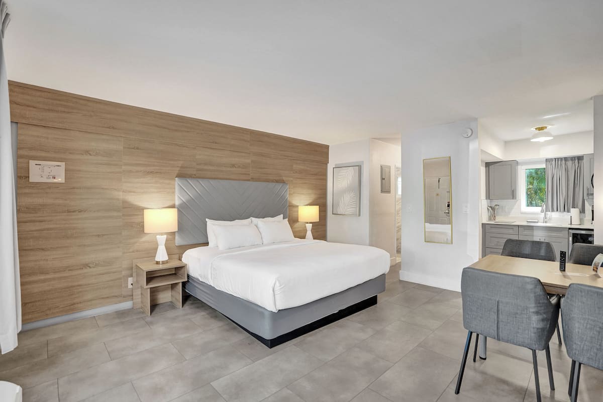 A modern hotel room with a large bed, bedside tables, a desk with chairs, a bathroom, and a kitchenette.