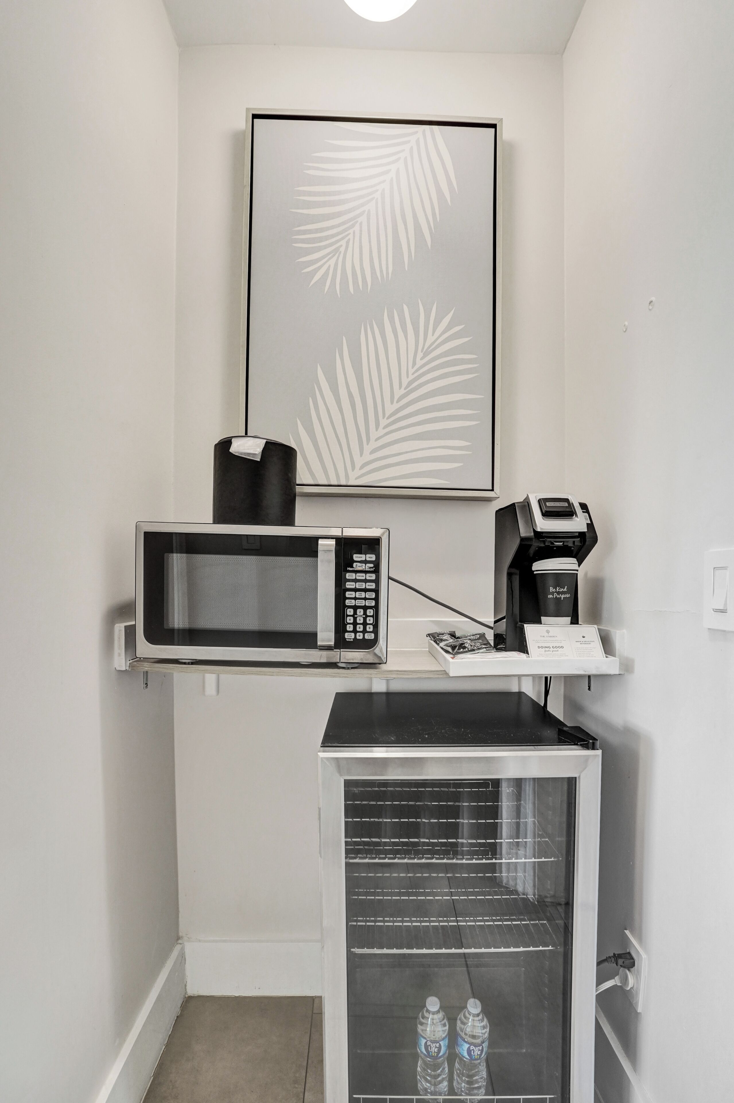 Una kitchenette con microondas, cafetera y un mini refrigerador que contiene botellas de agua.