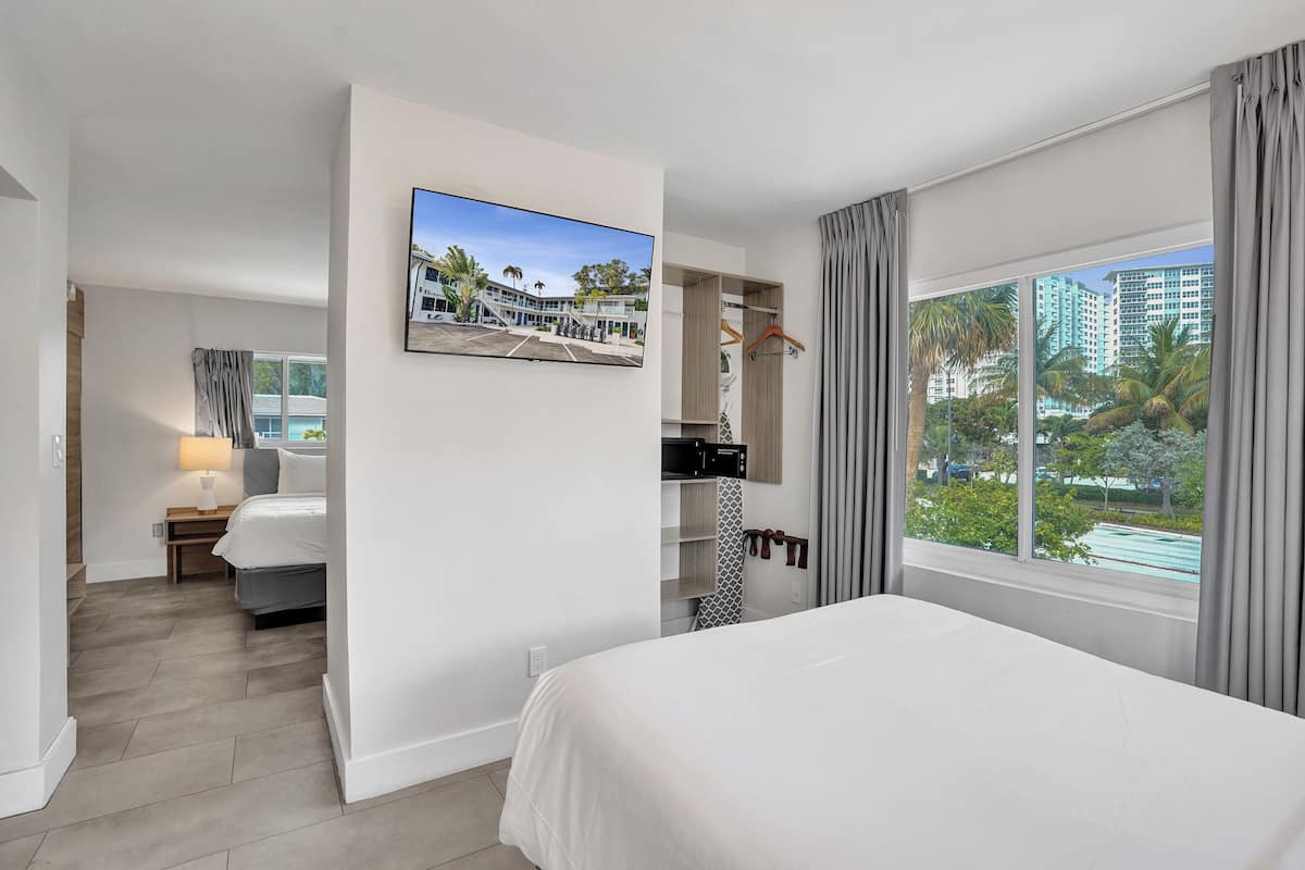 A hotel room with a bed, a TV mounted on the wall, a window with curtains, and a view of buildings and greenery outside.