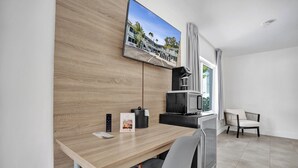 Una habitación de hotel moderna con escritorio de madera, televisor de pantalla plana montado en la pared, kitchenette, ventana con cortinas y un área de descanso con una silla.