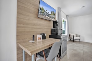 Una habitación de hotel moderna con escritorio de madera, televisor de pantalla plana montado en la pared, kitchenette, ventana con cortinas y un área de descanso con una silla.
