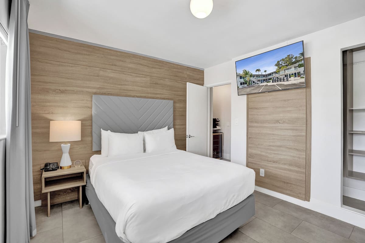 A hotel room with a bed, a nightstand, a lamp, a TV, and a view of a building outside.