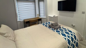Een slaapkamer met een bed, een bureau, een stoel, een televisie en twee ramen met jaloezieën.