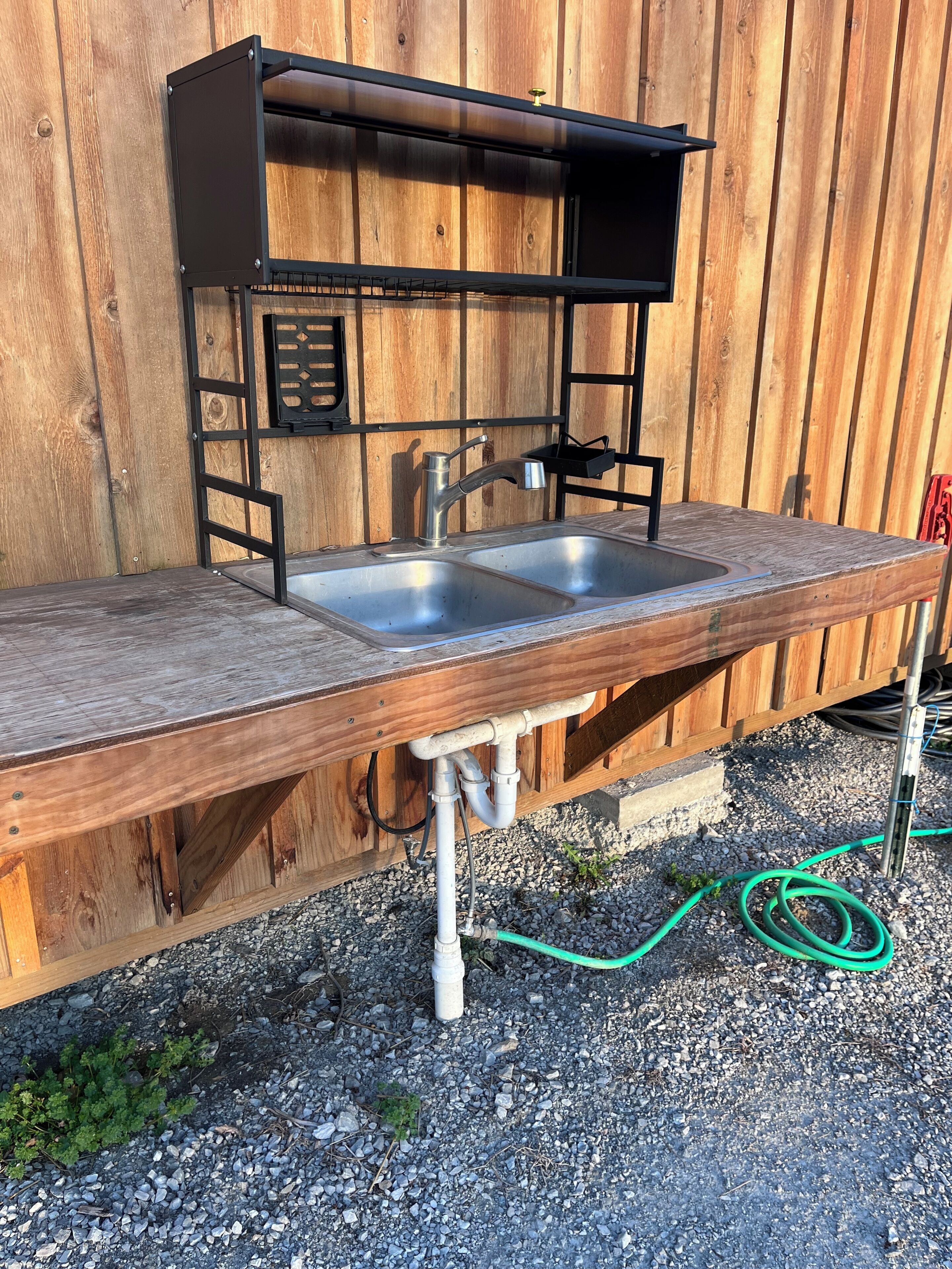 An outdoor kitchen with a metal sink and a wooden countertop, set against a wooden fence.