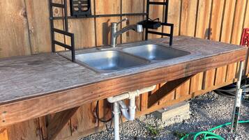 An outdoor kitchen with a metal sink and a wooden countertop, set against a wooden fence.