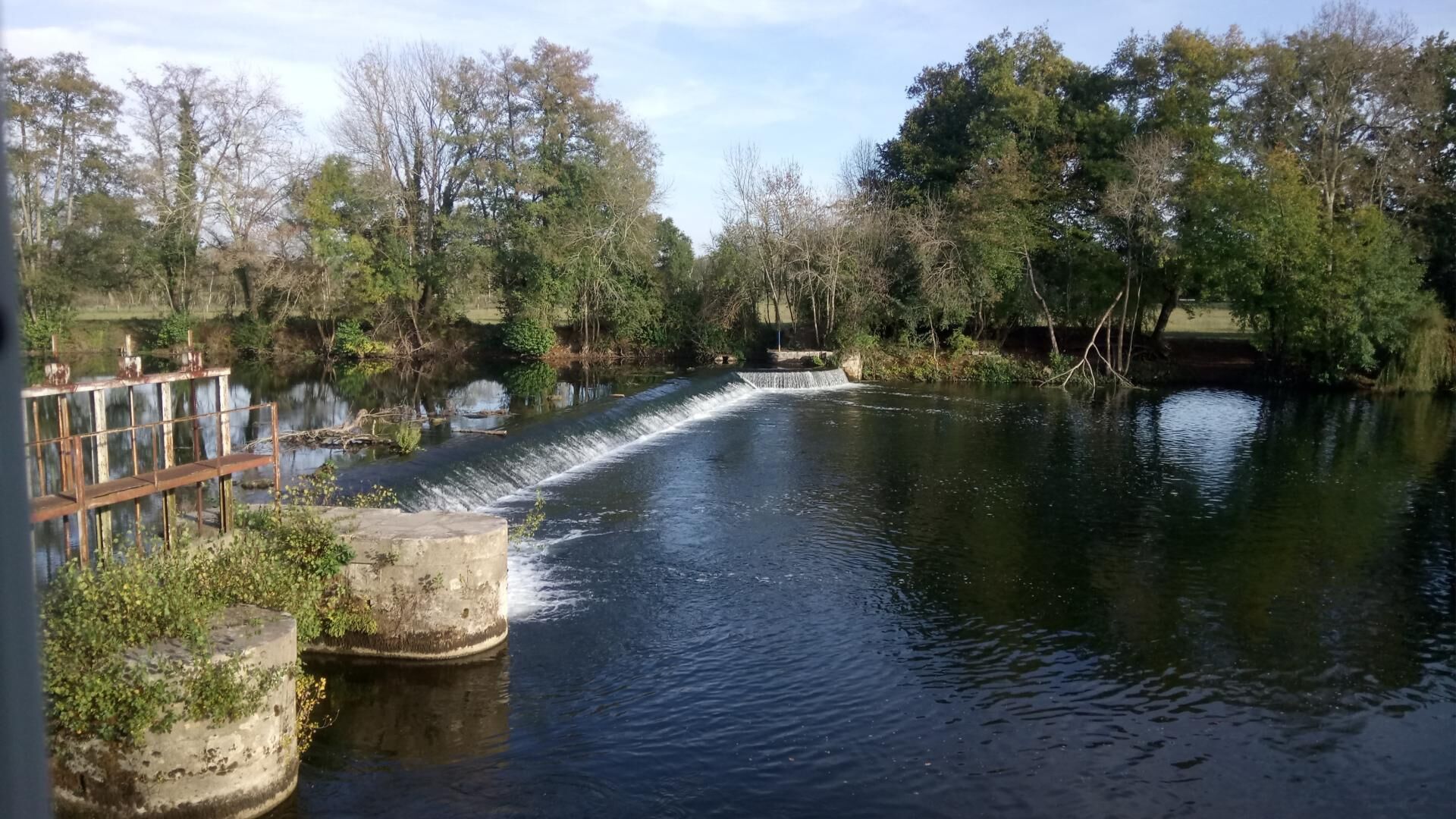 Um rio calmo com uma pequena cascata, rodeado por árvores e uma ponte de madeira.