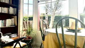 A room with a bookshelf, a dining table with a chair, a potted plant, and a view of the outdoors. - Charme d'Arès - ARES - Gîte (ARES)