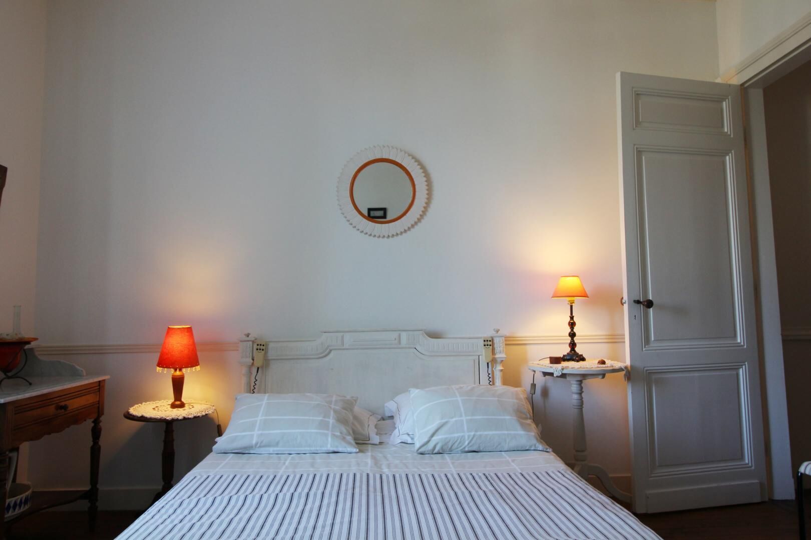 A hotel room with a bed, two bedside lamps, a round mirror, and a door.