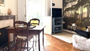 A living room with a wooden dining table, chairs, a TV stand with a flat-screen TV, a sofa, and a large painting of trees. - Charme d'Arès - ARES - Gîte (ARES)