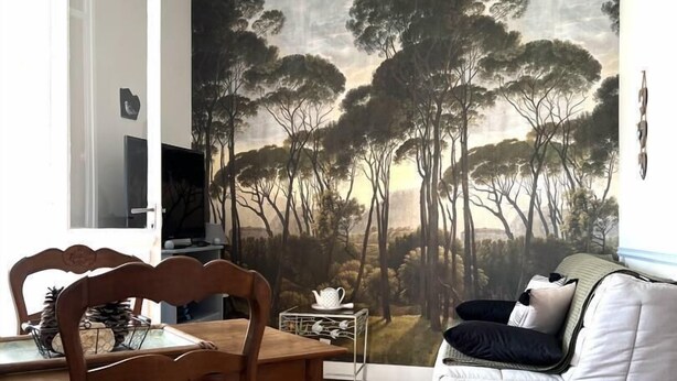 A dining area with a wooden table and chairs, a white sofa, and a large painting of trees on the wall. - Charme d'Arès - ARES - Gîte (ARES)