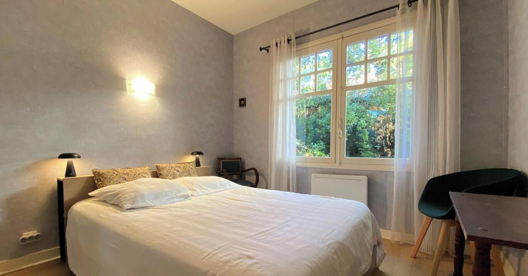 A bedroom with a bed, bedside lamps, a chair, a window with curtains, and a view of greenery outside.