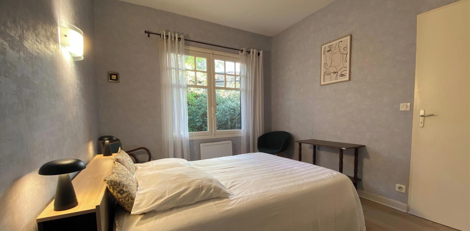 Ein Schlafzimmer mit einem Bett, einem Schreibtisch, einem Sessel, einer Lampe und einem Fenster mit Vorhängen.