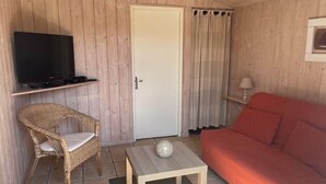 Ein Zimmer mit Fernseher auf einer Ablage, ein Korbstuhl, ein rotes Sofa, ein kleiner Tisch mit einer Dekokugel und eine Tür mit Vorhängen.