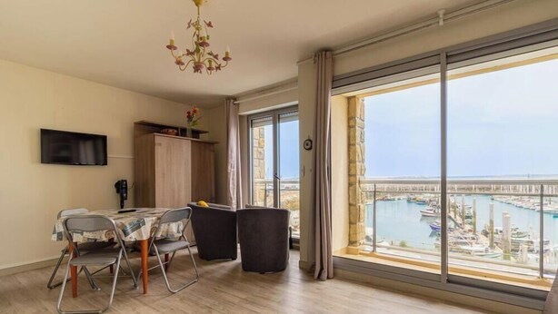 Une pièce comprenant une table à manger, des chaises, un téléviseur, un lustre et une grande fenêtre donnant sur une marina.
