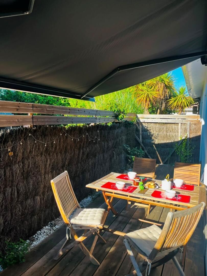 An outdoor patio with a table set for two, surrounded by wooden chairs, a thatched wall, and a wooden deck.