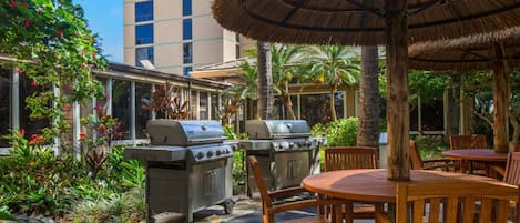 Eine Terrasse im Freien mit Grill, Holztisch und Stühlen, umgeben von Grünpflanzen und einem Gebäude im Hintergrund.
