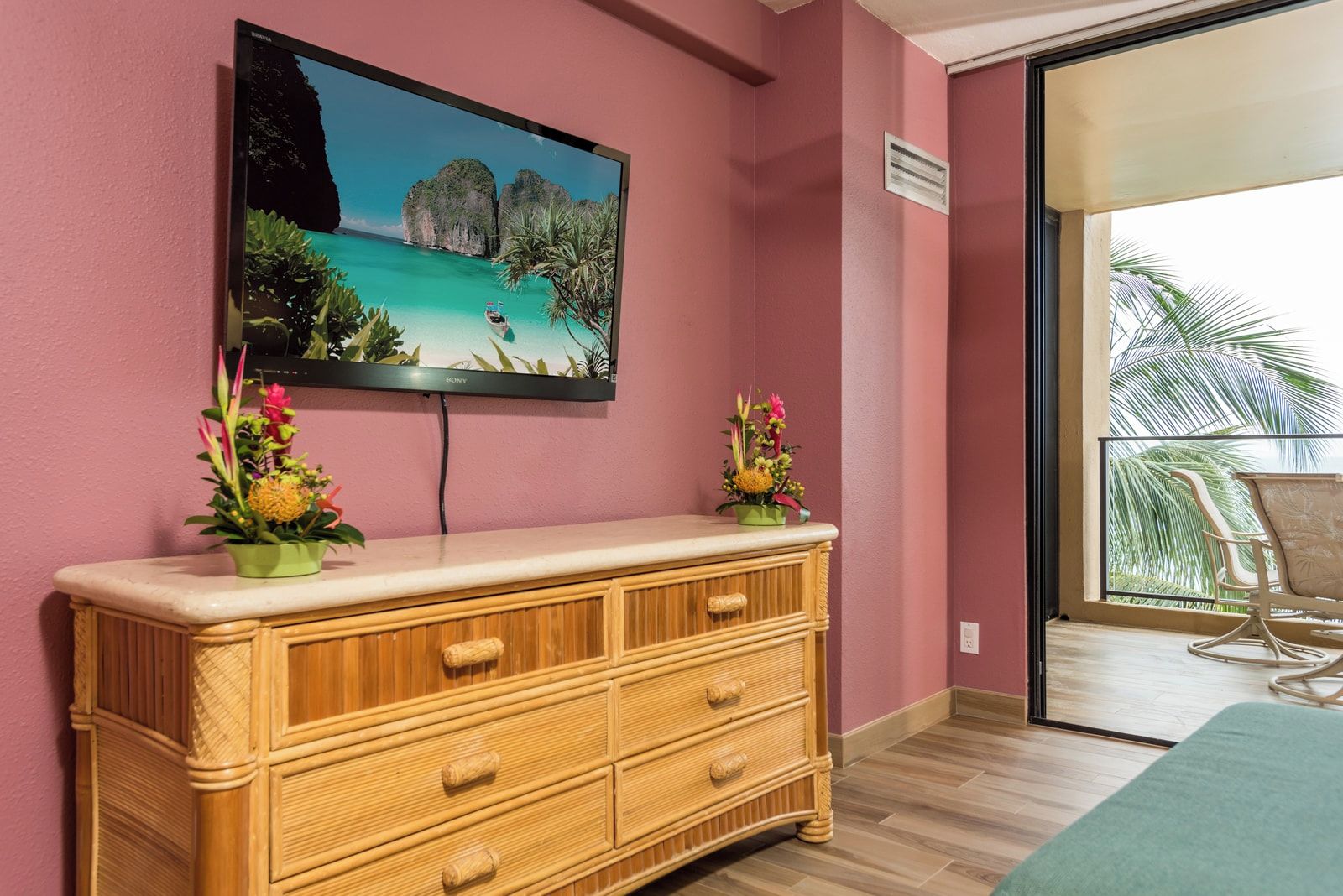 Una habitación con un televisor en la pared, un aparador de madera, dos arreglos florales y vistas a palmeras a través de un ventanal.