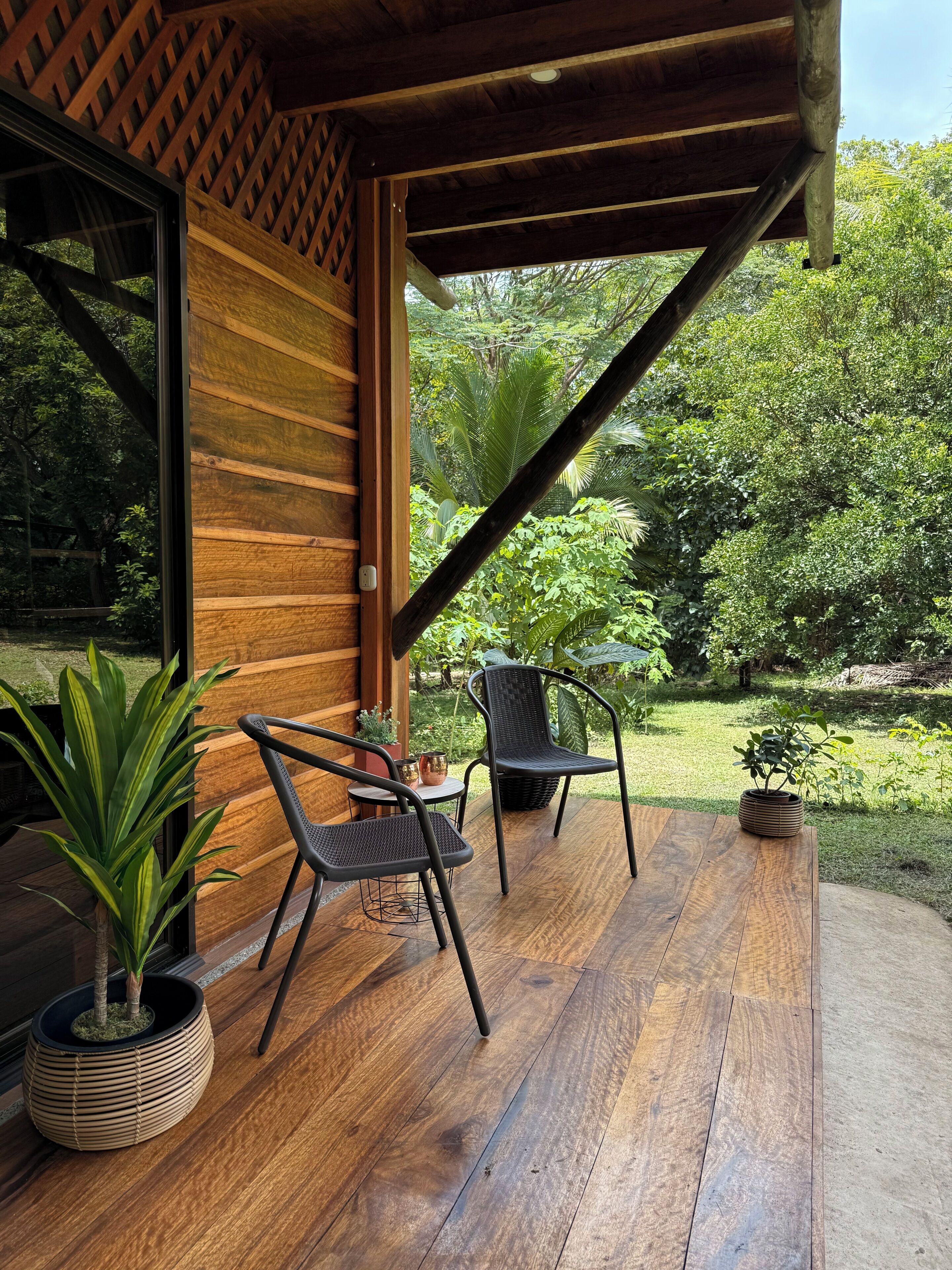 Una terraza de madera con dos sillas y una mesa, rodeada de exuberante follaje y un muro de madera.