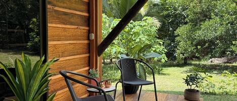 Una terraza de madera con dos sillas y una mesa, rodeada de exuberante follaje y un muro de madera.