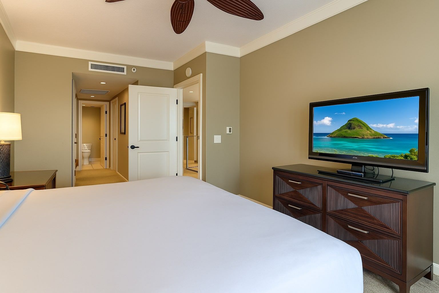 A hotel room with a bed, a TV on a wooden cabinet, a door leading to a bathroom, and a ceiling fan.