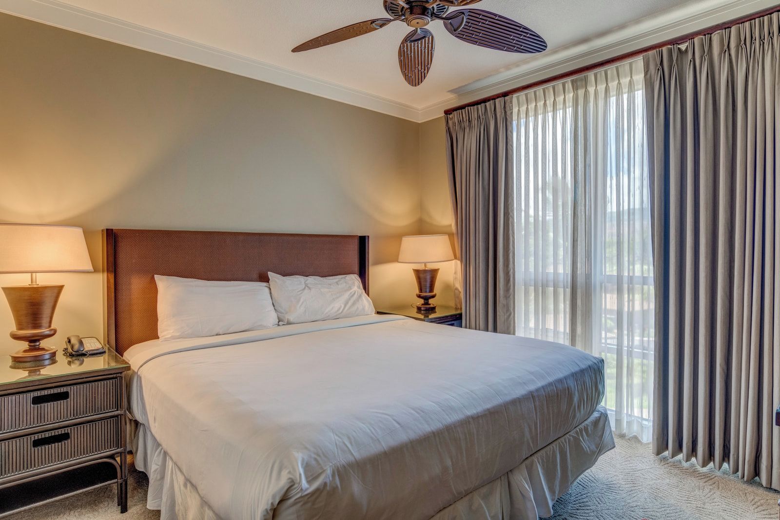 A hotel room with a large bed, two bedside tables with lamps, a ceiling fan, and a view of the outdoors through curtains.