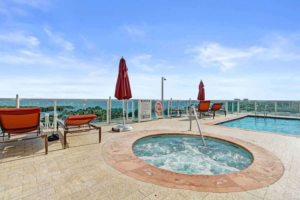 A rooftop pool area with lounge chairs and umbrellas, a hot tub, and a view of the ocean.