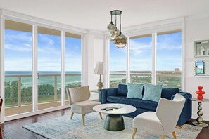 A spacious living room with a large window overlooking the ocean, a blue sofa, two armchairs, a round coffee table, and a hanging light fixture. - Bay View 2BR | Rooftop Pool, Walk to CocoWalk (Miami)