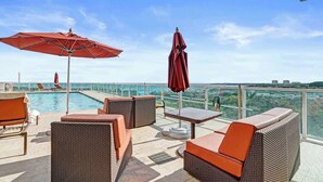 Una terraza en la azotea con mobiliario de mimbre, una piscina y una barandilla de cristal con vistas a un paisaje urbano.
