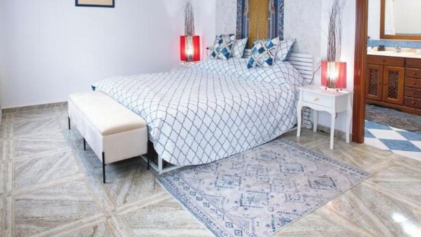 Une chambre à coucher comprenant un lit, des tables de chevet, une commode, un banc, un tapis à motifs, un mur agrémenté d’une arche décorative et un tableau encadré.
