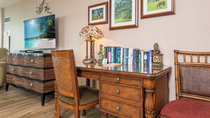 Ein Zimmer mit einem Holztisch, Büchern, einer Lampe, einem Fernseher und gerahmten Bildern an der Wand.