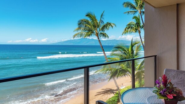 Ein Balkon mit Blick auf das Meer und ein Berg, ein kleiner Tisch mit einer Pflanze und ein Stuhl.