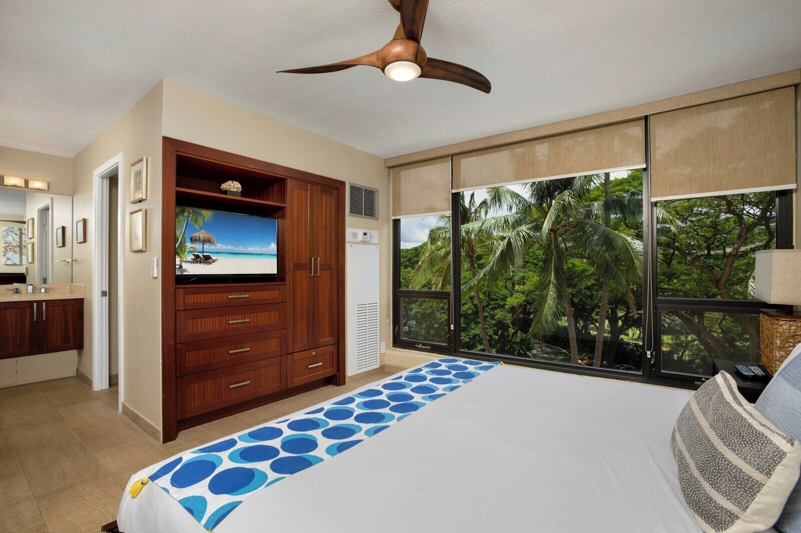 A bedroom with a bed, a television on a wooden cabinet, a ceiling fan, and a large window with a view of palm trees.