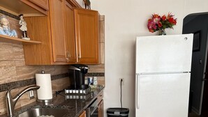 Een keuken met granieten aanrechtbladen, houten keukenkastjes, een roestvrijstalen spoelbak, een koffiezetapparaat, een witte koelkast en een vuilnisbak.