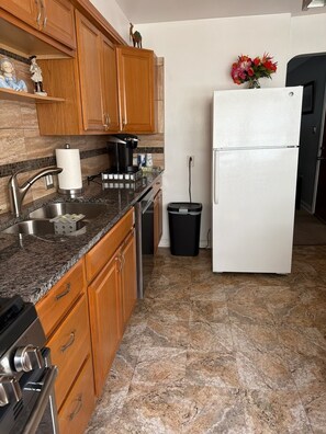 Una cocina con encimeras de granito, gabinetes de madera, un lavamanos de acero inoxidable, una cafetera, un refrigerador blanco y un cubo de basura.
