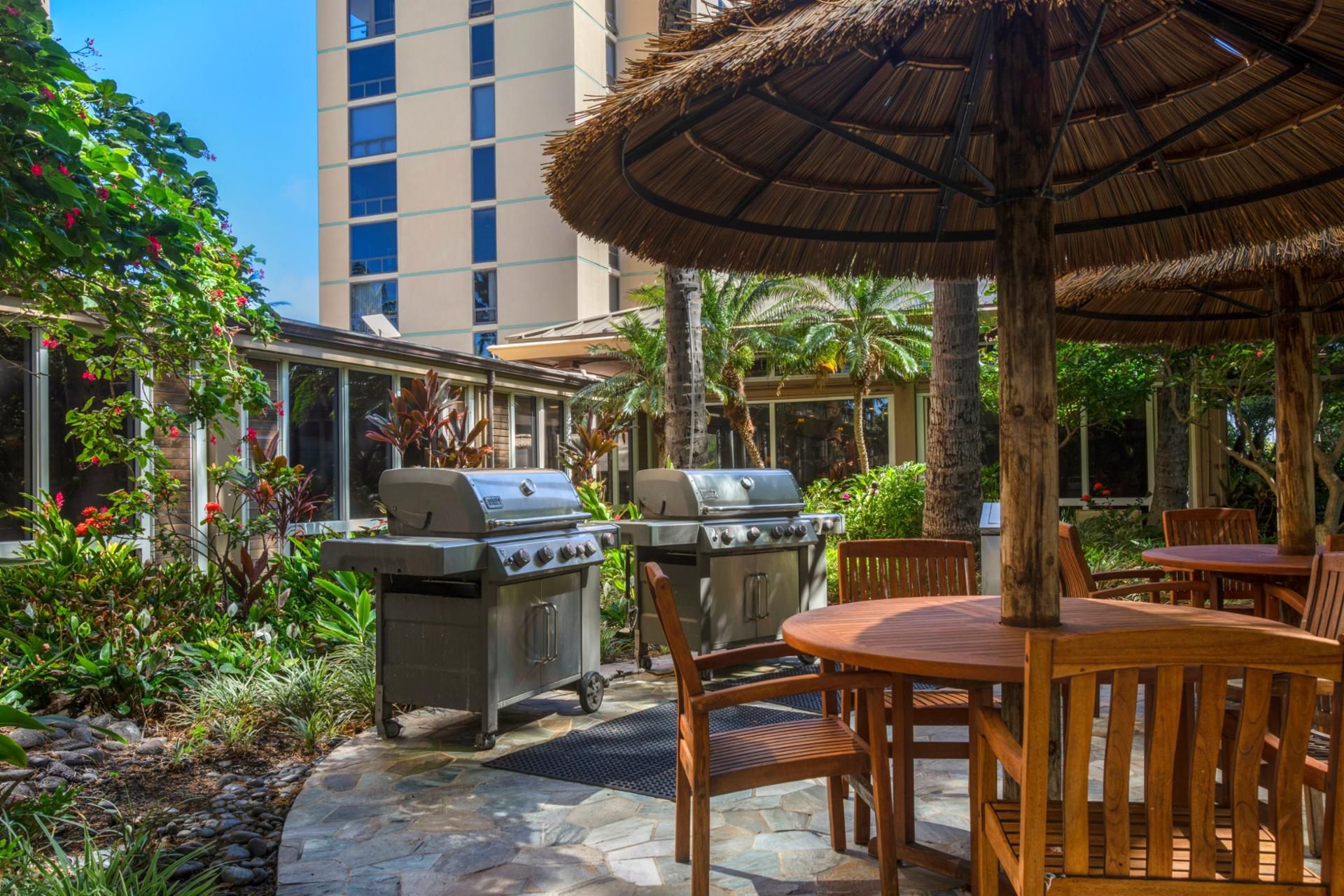 An outdoor patio with a grill, wooden table, and chairs, surrounded by greenery and a building in the background.