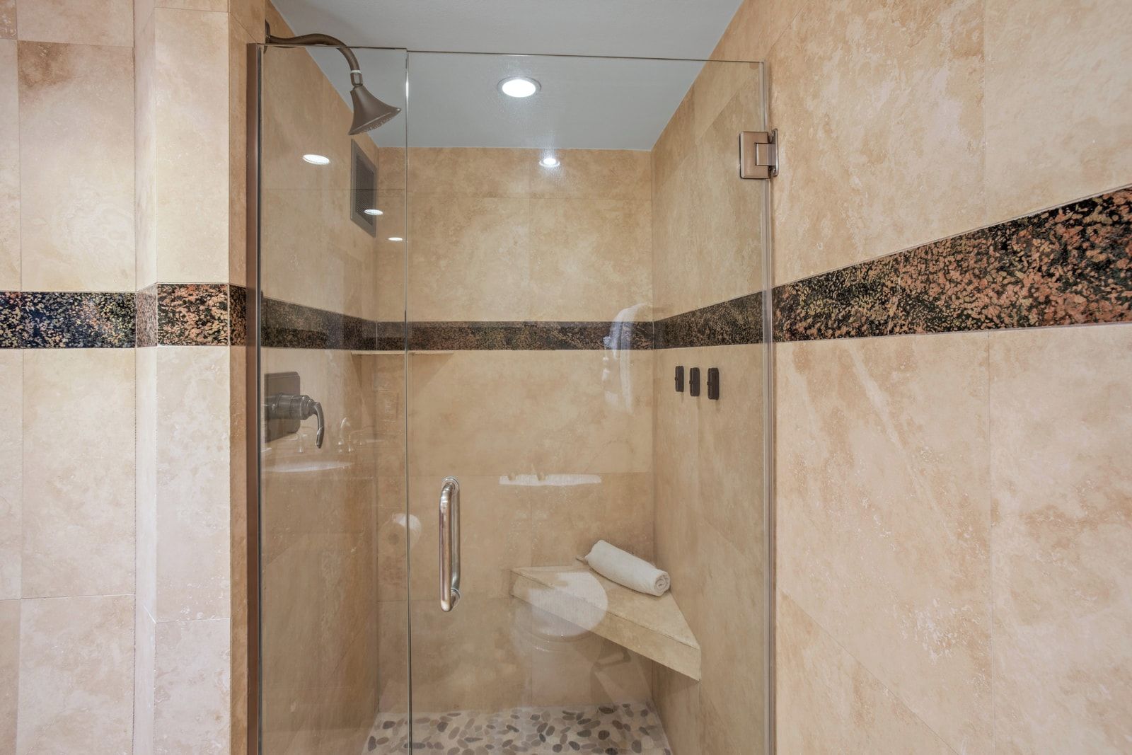 Une douche fermée par des parois de verre, dotée d’un pommeau de douche à main, d’un porte-serviettes et d’un mur recouvert de carreaux de marbre.