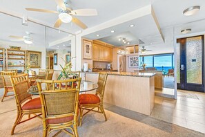 Amplia sala de estar con mesa de comedor, sillas de mimbre, ventilador de techo y cocina con alacenas de madera y un ventanal grande.