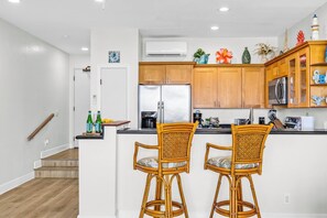 A modern kitchen with wooden cabinets, a central island with two wicker chairs, and a stainless steel refrigerator. - Napili Point | Oceanfront 4 BR, Sleeps 14 | Car Incl. w/ 6+ Nights | NAP ML-3897 by KBM (Lahaina)