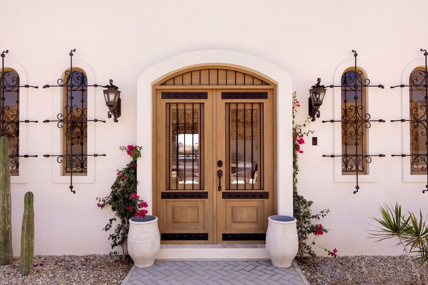 Une porte en bois à deux battants, surmontée d’une arche décorative, et flanquée de deux grands jardinières et de deux lanternes fixées au mur.