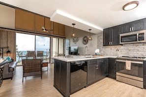 A modern kitchen with dark cabinetry, a large island, stainless steel appliances, and a view to the outside. - Napili Point | Oceanfront 5 BR, Sleeps 14 | Car Incl. w/ 6+ Nights | NAP ML-3909 by KBM (Lahaina)