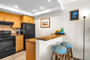 A kitchen with wooden cabinets, a black refrigerator, a granite countertop, and two blue stools. - Napili Point | Oceanfront 3 BR, Sleeps 12 | Car Incl. w/ 6+ Nights | NAP ML-3924 by KBM (Lahaina)