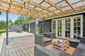 An outdoor patio with wooden decking, wicker furniture, and a glass-enclosed area with a wooden ceiling. - Cozy holiday home in peaceful Bredfjed (Rødby)