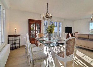 A dining area with a glass table, white chairs, a chandelier, and a sofa in the background. - A l'abri de Flo - CANCALE (CANCALE)