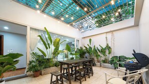 A covered patio with a wooden table and chairs, surrounded by potted plants and a grill. - BMC BUILDING (Ho Chi Minh City)